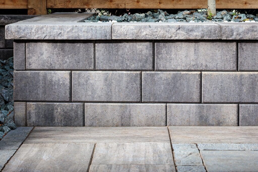 Close-up of a modern grey concrete block retaining wall with chiseled cap stones next to large patio pavers - Yellow Brick Road Pavers & Landscapes LLC