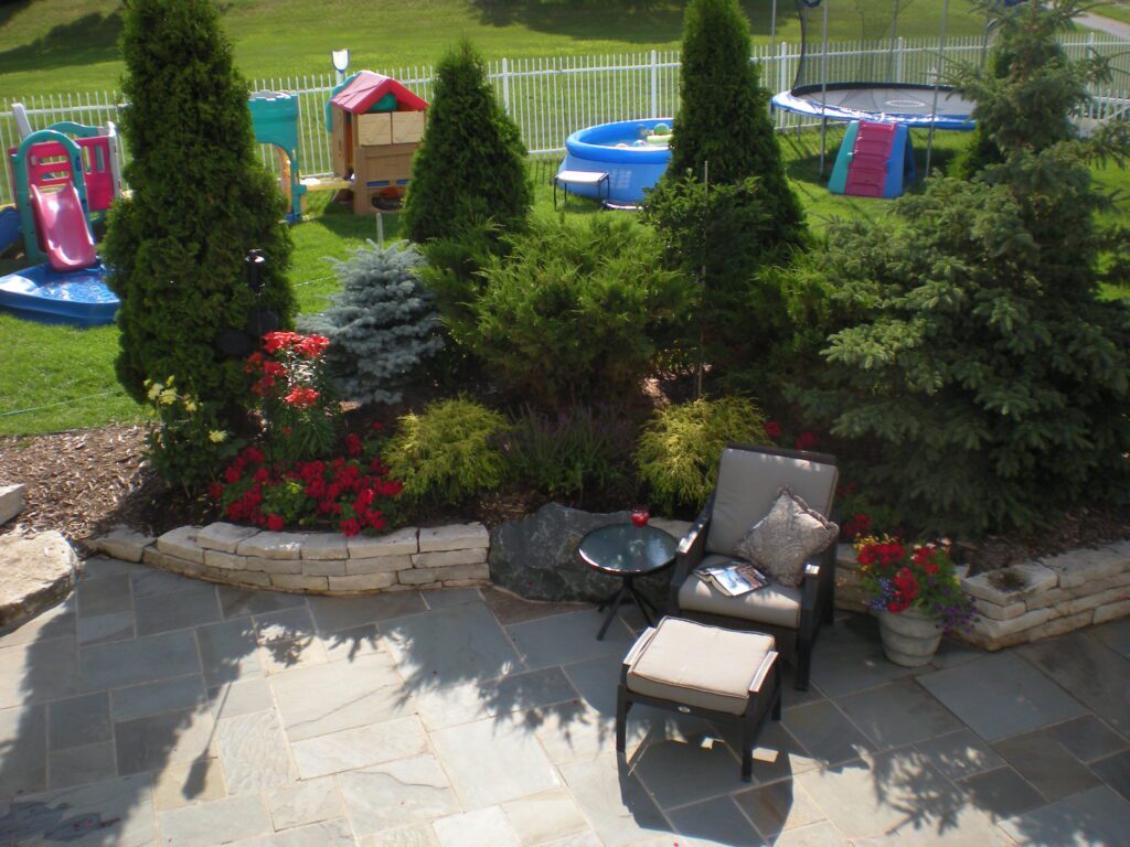 Lush garden scene featuring a cozy seating area, surrounded by colorful flowers, with children's play equipment and a trampoline in the background - Yellow Brick Road Pavers & Landscapes LLC