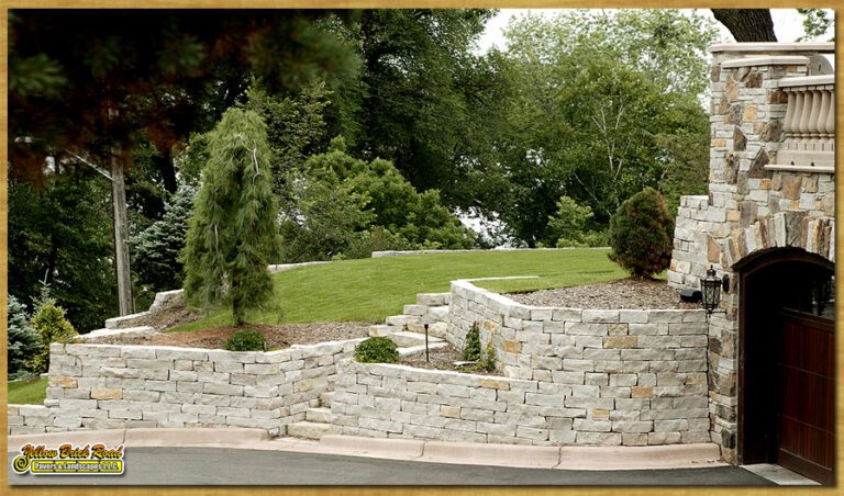 A landscaped stone wall curves up a sloped yard, bordered by lush greenery, enhancing the home's exterior and outdoor appeal - Yellow Brick Road Pavers & Landscapes LLC