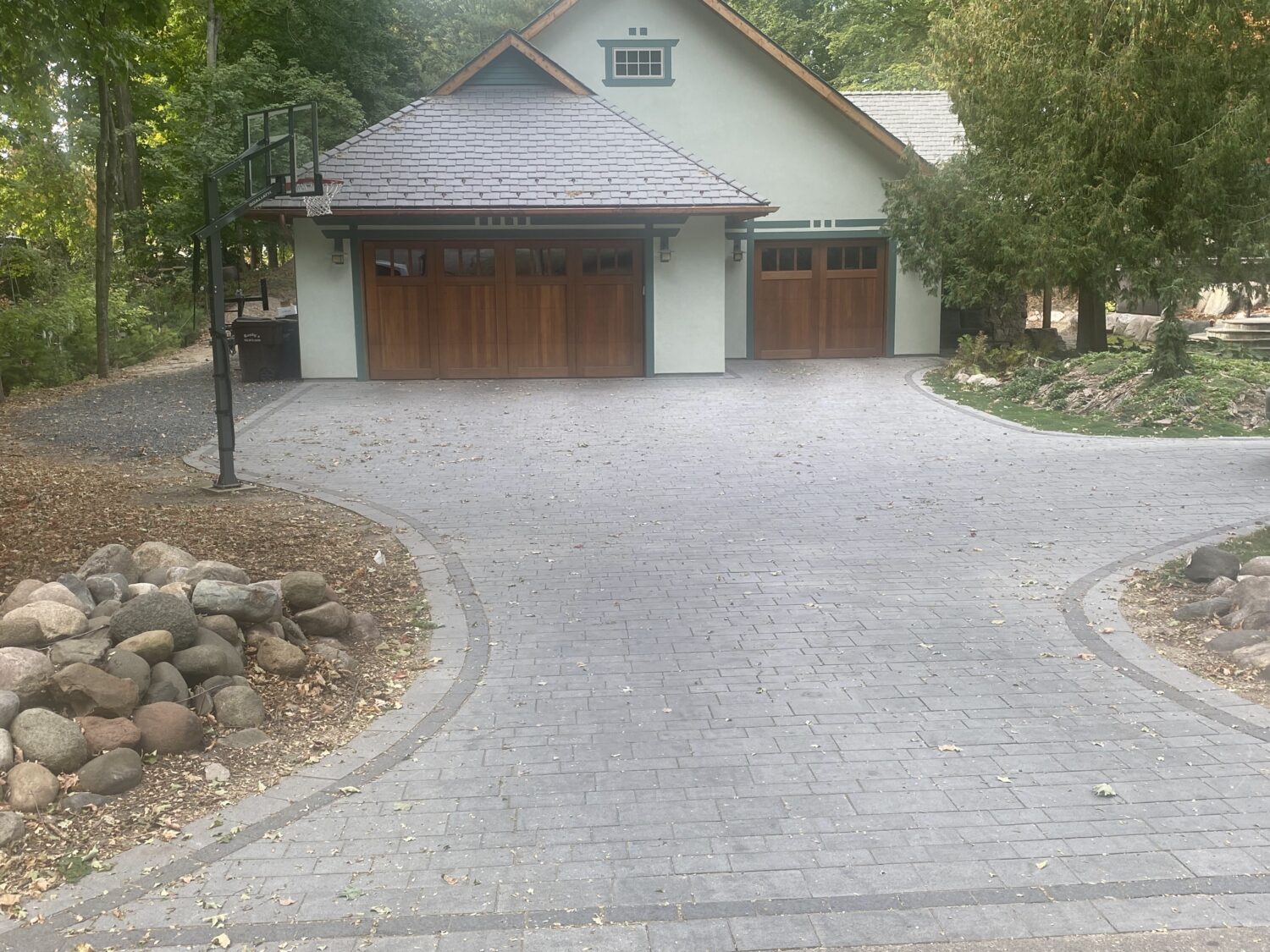 A modern house with wooden garage doors and a paved driveway, featuring a basketball hoop nearby and surrounded by trees and landscaping.