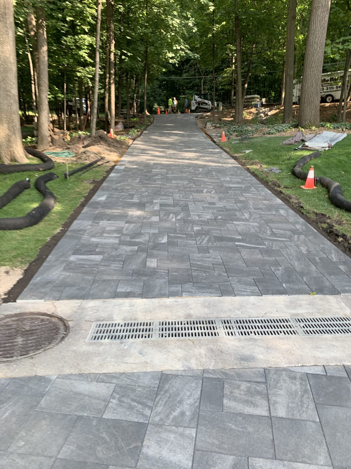 Newly installed gray paver driveway winds through a wooded area, flanked by construction markers and equipment, indicating ongoing landscaping work.