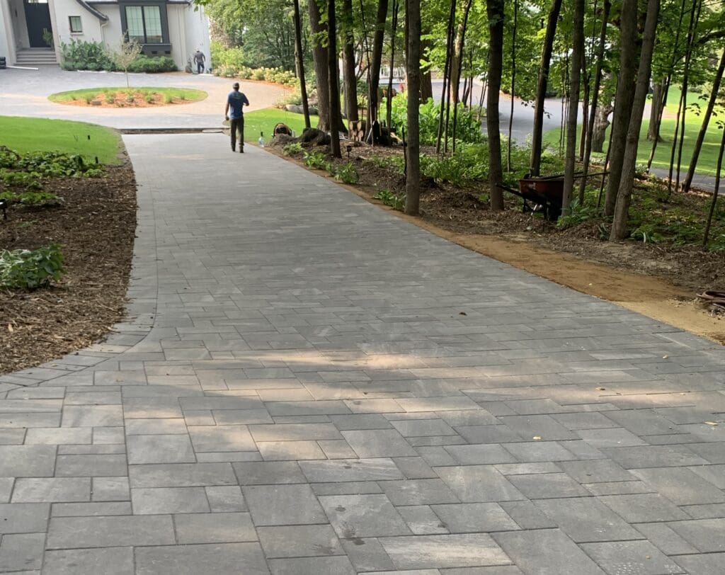A paved pathway winds through a landscaped area, leading toward a house. A person walks along the path, showcasing outdoor design and accessibility - Yellow Brick Road Pavers & Landscapes LLC