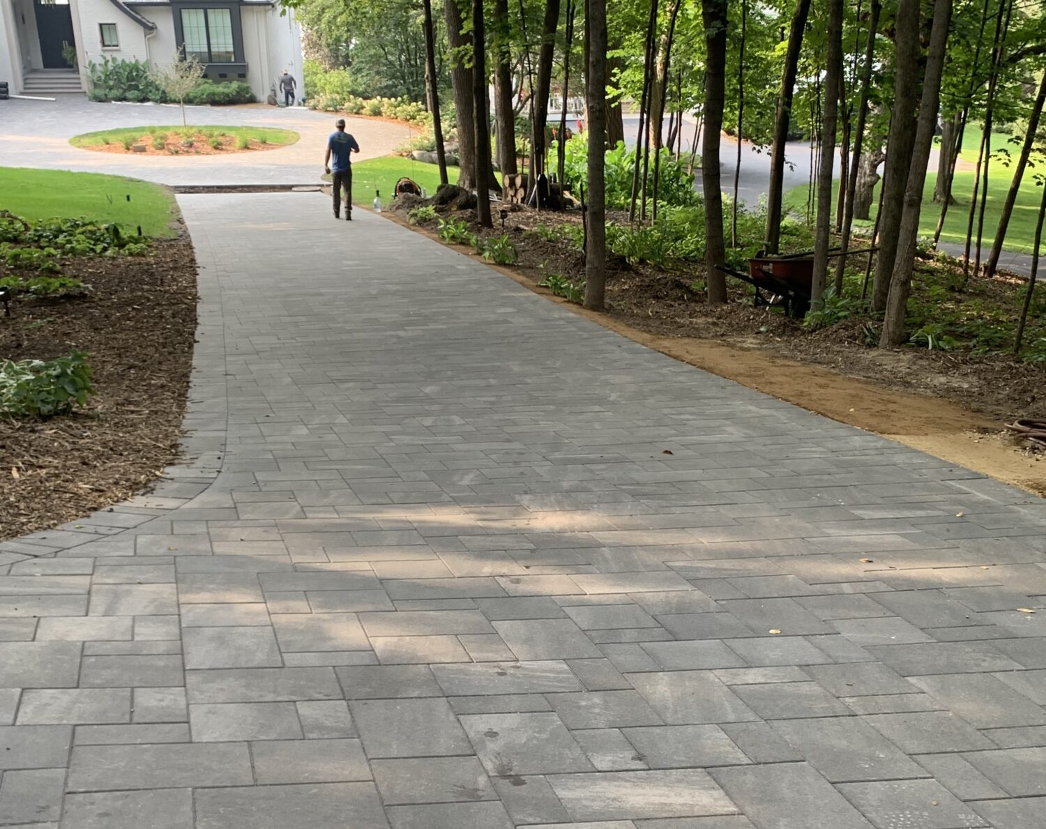 A paved pathway winds through a landscaped area, leading toward a house. A person walks along the path, showcasing outdoor design and accessibility.