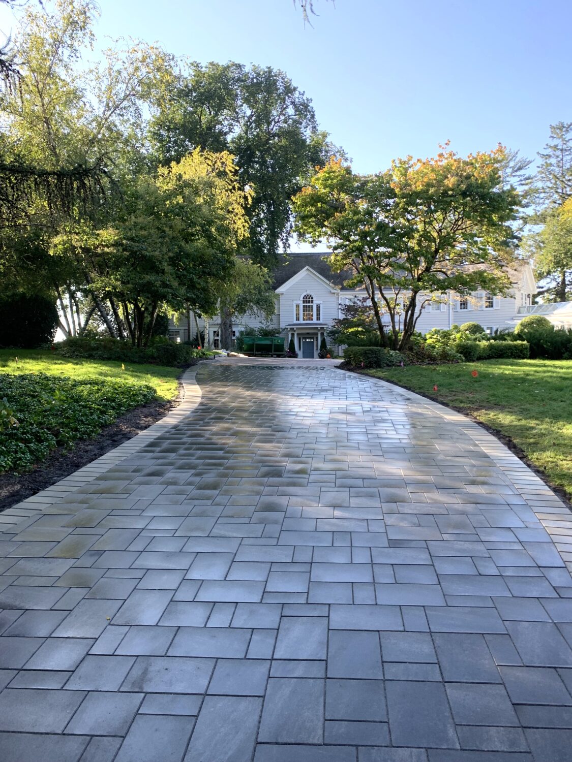 Paved walkway leading to a large, light-colored house surrounded by trees and greenery, highlighting a serene residential setting.