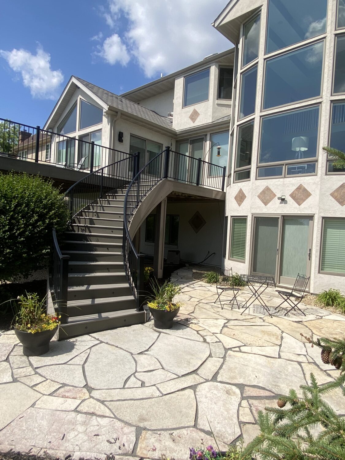 Modern two-story home with large windows, featuring a curved staircase leading to a stone patio area with planters and outdoor seating.