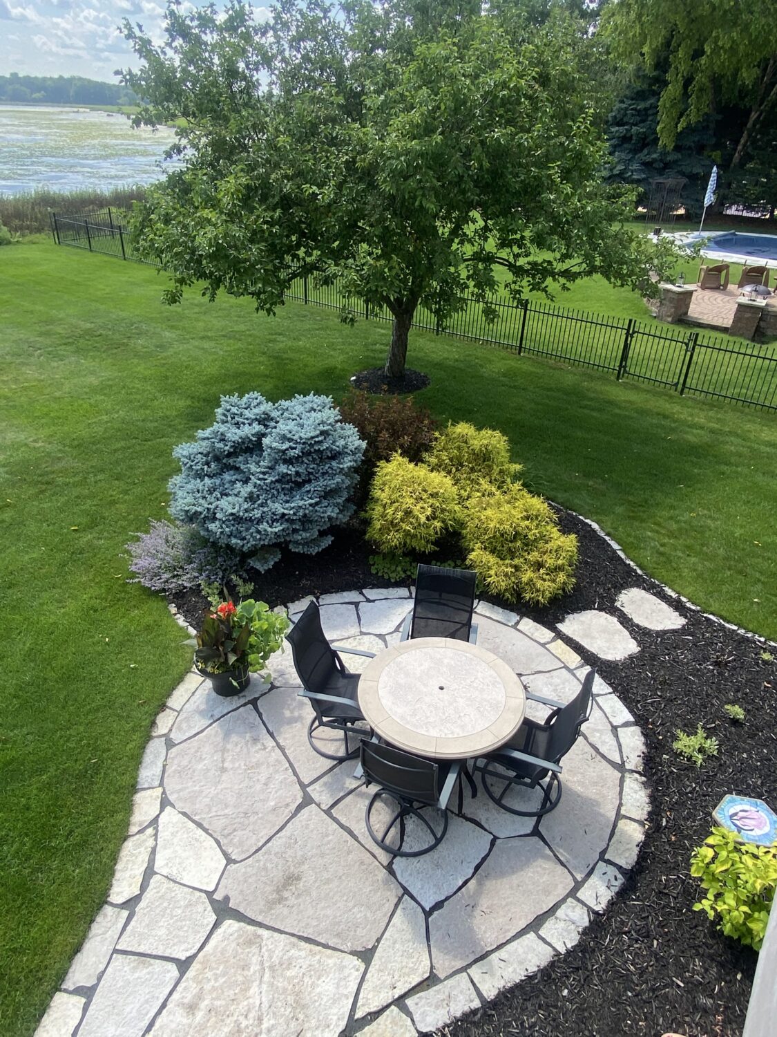A stone patio with a circular table and six black chairs surrounded by lush greenery, overlooking a serene lake in the background.