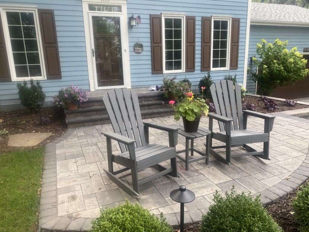 Two gray Adirondack chairs and a small table on a paved patio, adorned with colorful flowers and set against a blue house exterior - Yellow Brick Road Pavers & Landscapes LLC