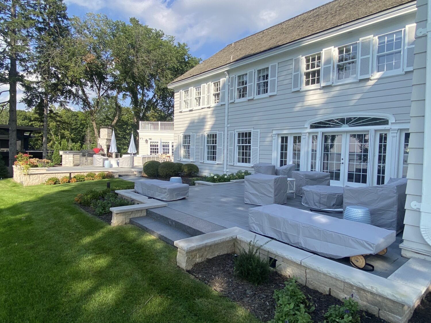 Spacious patio area featuring covered outdoor furniture and a grill, surrounded by lush landscaping and a two-story house in a tranquil setting.