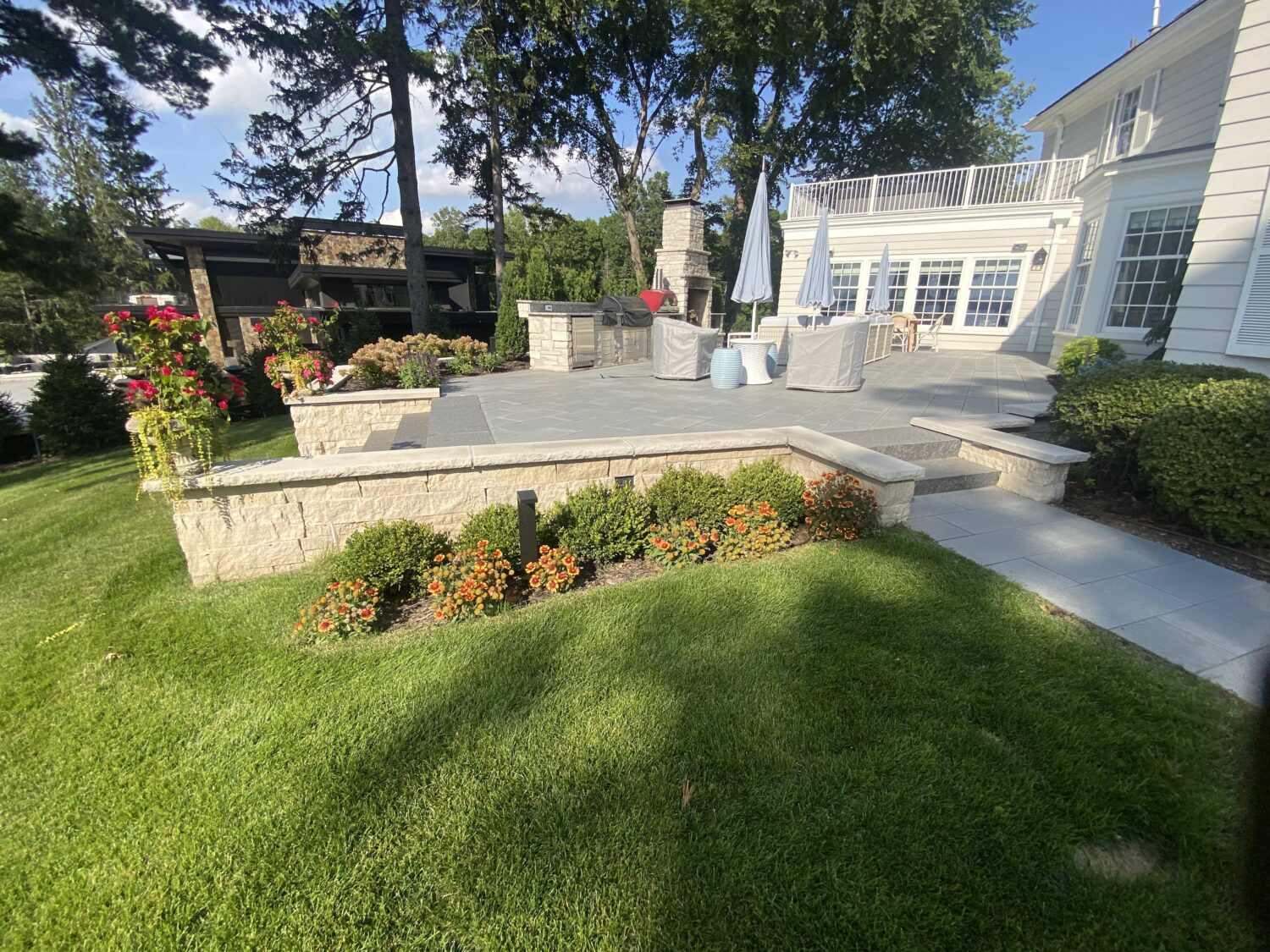 A landscaped backyard features stone steps leading to a spacious patio with outdoor furniture, surrounded by colorful flowers and greenery.