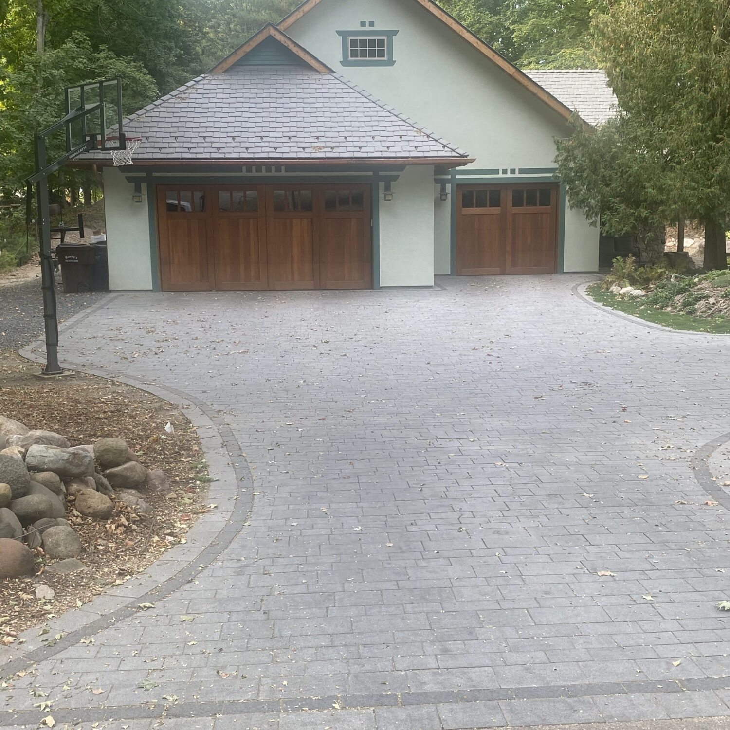A modern house with wooden garage doors and a paved driveway, featuring a basketball hoop nearby and surrounded by trees and landscaping - Yellow Brick Road Pavers & Landscapes LLC